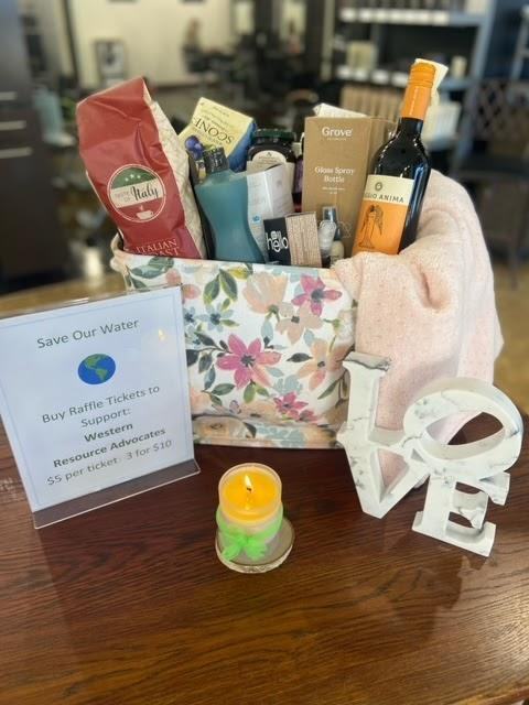 A floral gift basket with assorted items such as a wine bottle, coffee, snacks, and toiletries is on display. The basket includes products for hair and skin care. A sign nearby promotes a beautiful raffle for Western Resource Advocates, with ticket prices listed.