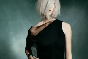 Person with a chin-length bob haircut in platinum blonde, hair covering one eye, wearing a black asymmetrical dress, stands against a dark background—capturing an edgy spring vibe.