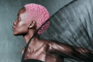A person with a short pink bob haircut and dark skin poses in profile against a gray background, wearing a sheer black garment that fans out behind them—a bold look perfect for spring.