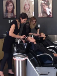 4-Centre-Salon-Arvada-After -(6) Two hairstylists wash the hair of two women reclining in salon chairs, with hair color posters visible on the wall behind them.