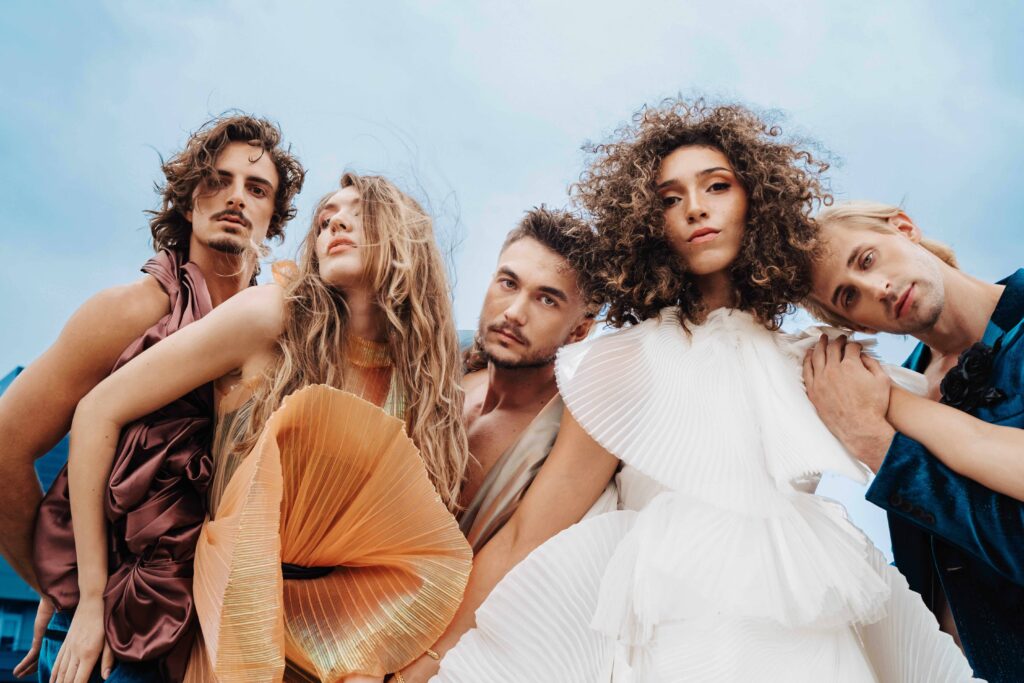 CentreSalons_Denver-22 Five people wearing avant-garde, pleated fashion garments stand closely together, posing outdoors against a blue sky, looking directly at the camera.