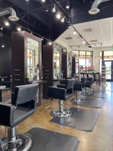 IMG_6461 Interior of a modern hair salon with multiple black styling chairs, stations with mirrors, and shelves displaying hair products. The room is well-lit with natural and artificial light.