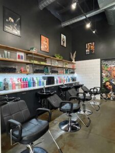 IMG_6463 Three black salon chairs with sinks are lined up in front of shelves holding hair products and towels, with framed photos and posters on the black and white walls.