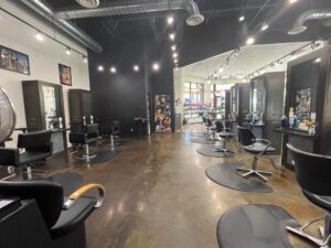 IMG_6467 Spacious modern hair salon with empty black styling chairs and mirrors, polished concrete floor, and hair products displayed on shelves in the background.