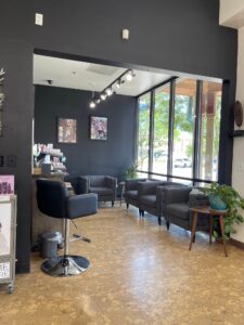 IMG_6468 Modern salon waiting area with four gray armchairs, large windows, a plant, artwork on the wall, and a styling chair in the foreground. Natural light fills the space.