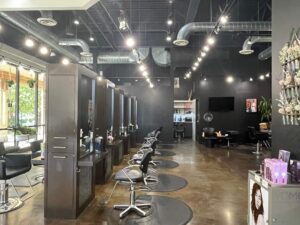 IMG_6472 Modern hair salon interior with multiple styling stations, black chairs, large mirrors, bright overhead lighting, and shelves with hair products.