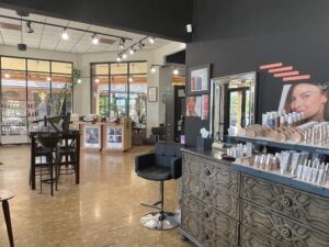 IMG_6473 A modern salon interior with styling chairs, a display of makeup products, large mirrors, and natural light from windows.