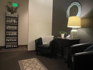 IMG_6475 A small waiting area with two black chairs, a side table with a lamp and plant, a decorative mirror, and a shelf with products near an exit sign.