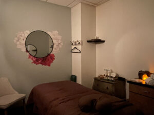 IMG_6480 A spa treatment room with a massage table, a chair, a round mirror with floral decor, shelves with products, and soft ambient lighting.