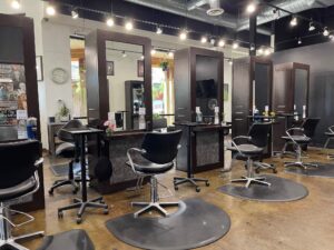 IMG_6483 A modern hair salon interior with empty black styling chairs, mirrors, and workstations under bright ceiling lights.