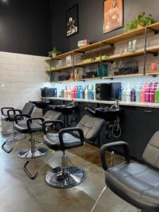 IMG_6485 A row of black salon chairs faces shampoo sinks, with shelves above holding hair products and decor in a modern, well-lit salon.