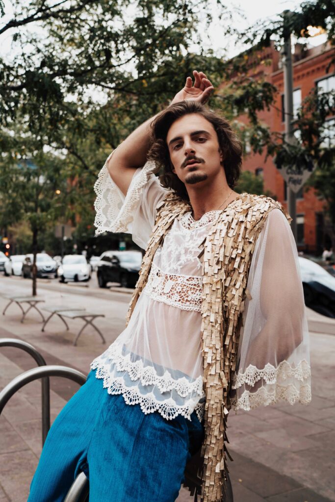 Salons_Denver-20 A person with long hair and a mustache poses confidently outdoors, wearing a sheer lace top, fringe vest, and blue pants, with trees and parked cars in the background.