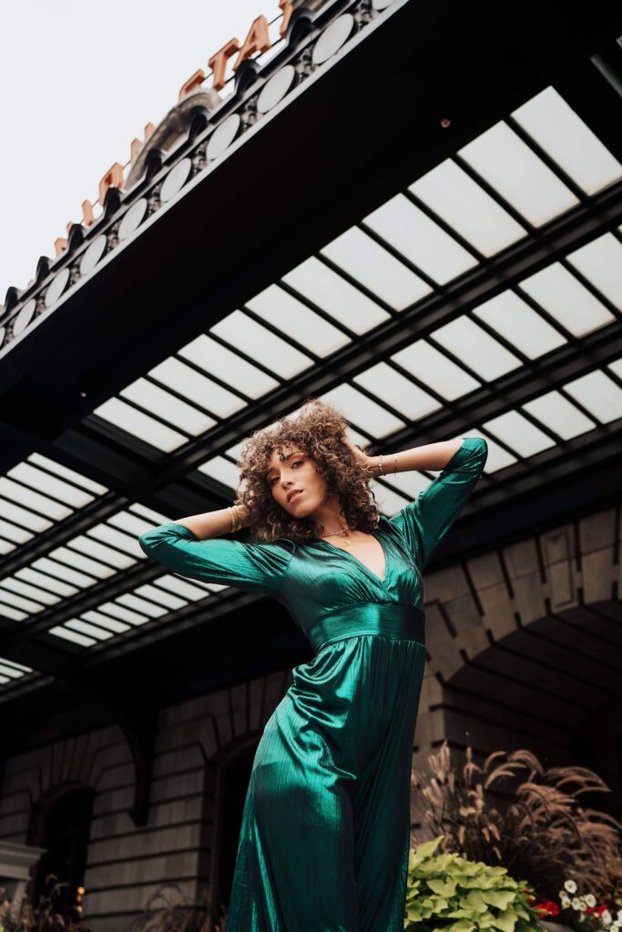 Salons_Denver-3 A person in a shiny green dress poses with hands behind their head in front of a building with a glass canopy and partial sign overhead.