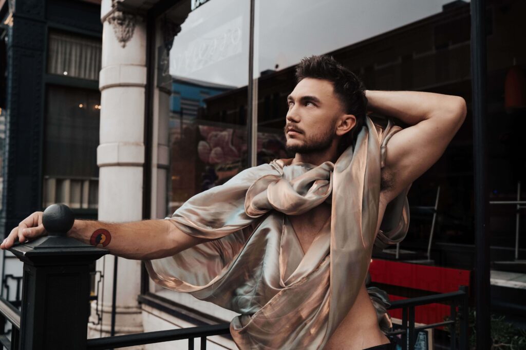 Salons_Denver-32 A man poses outdoors, leaning on a railing, wearing a sheer, draped fabric top with one arm behind his head and looking off into the distance.