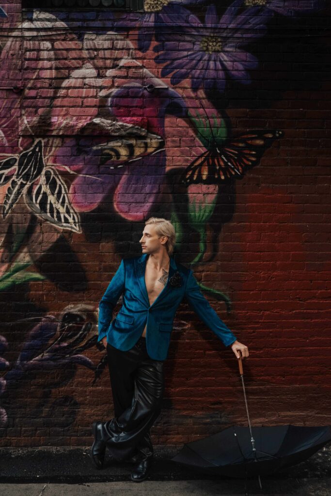 Salons_Denver-42 A person in a teal blazer and black pants leans against a mural-covered brick wall, holding a closed umbrella beside them.