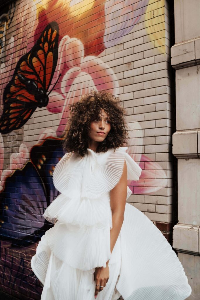 Salons_Denver-49 A woman in a tiered white dress stands in front of a brick wall with colorful butterfly and flower graffiti.