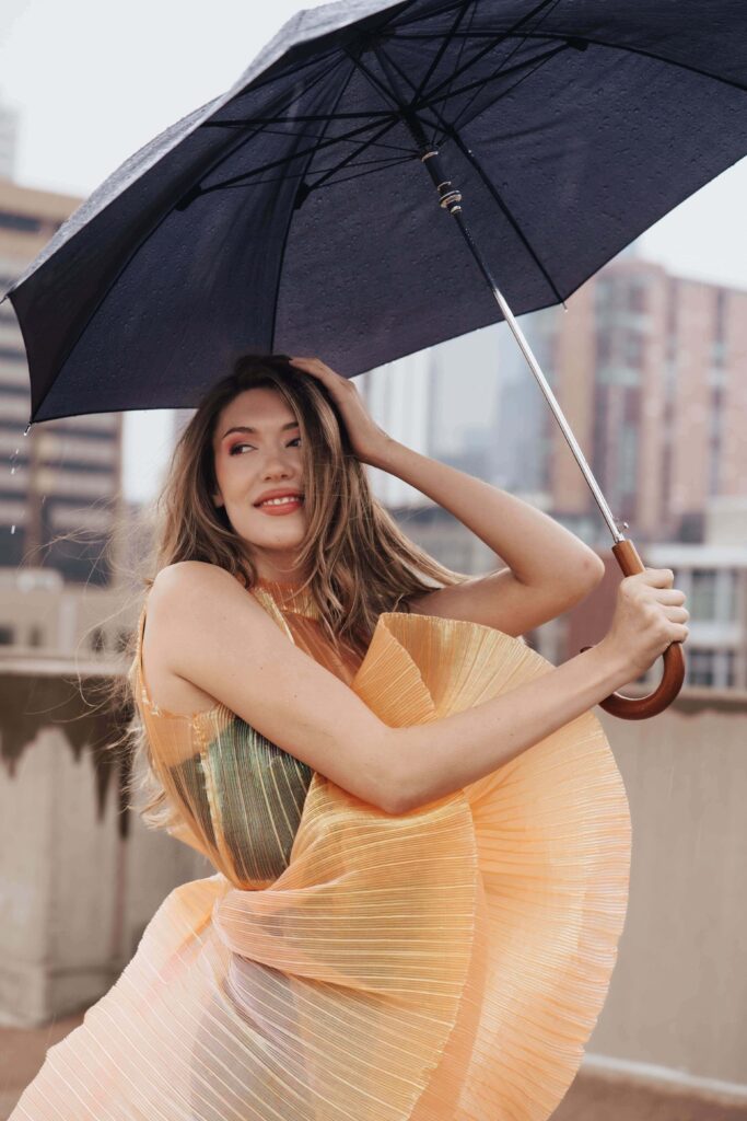 Salons_Denver-71 A woman in a pleated orange dress holds a black umbrella on a city rooftop in the rain, with buildings blurred in the background.