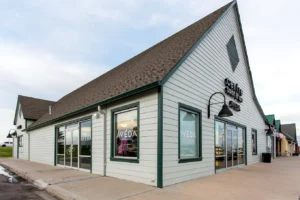 centresalon-arvada-3 A modern light-gray commercial building with large windows, labeled “Aveda” and “Castle Rock Salon,” set against a cloudy sky with a sidewalk in front.