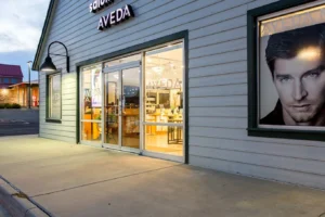 centresalon-arvada-39 Storefront of an Aveda salon with glass double doors, illuminated interior, and a large poster of a man's face in the window.