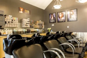 centresalon-arvada-62 Five black salon chairs are lined up in front of hair washing sinks, with shelves of hair products and posters on the wall in a modern salon setting.
