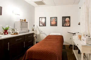 centresalon-arvada-74 A treatment room with a brown quilted bed, dark cabinets, medical equipment, and three framed photos on the wall. White walls and ceiling with a vase of flowers on the counter.