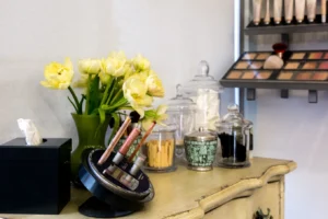 centresalon-arvada-78 A vanity table with makeup brushes, cosmetics, glass jars, a tissue box, and a vase of yellow tulips arranged on top.