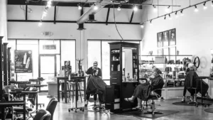 tiffany-plaza Black and white photo of a modern hair salon with two clients seated at stations, stylists working, and shelves stocked with products in the background.