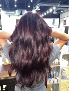 Person with long, wavy dark burgundy hair stands indoors with their back to the camera, holding their hair up with both hands in a well-lit salon setting.