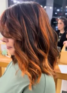 Woman with medium-length, wavy hair featuring dark brown and copper-orange highlights, standing indoors with blurred background and another person visible.