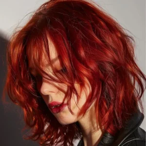 Person with wavy, shoulder-length bright red hair looking downward. They are wearing a black garment and are positioned against a neutral background.