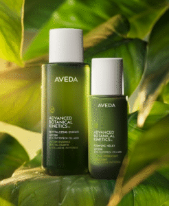 Two green Aveda Advanced Botanical Kinetics skincare bottles are displayed on green leaves, featuring Revitalizing Essence and Plumping Milky Lotion.