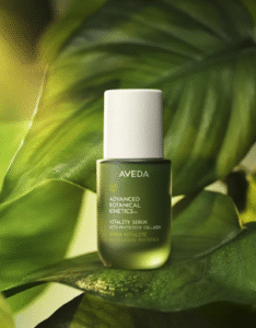 A green bottle of Aveda Advanced Botanical Kinetics Vitality Serum is displayed on a large leaf with soft natural lighting.